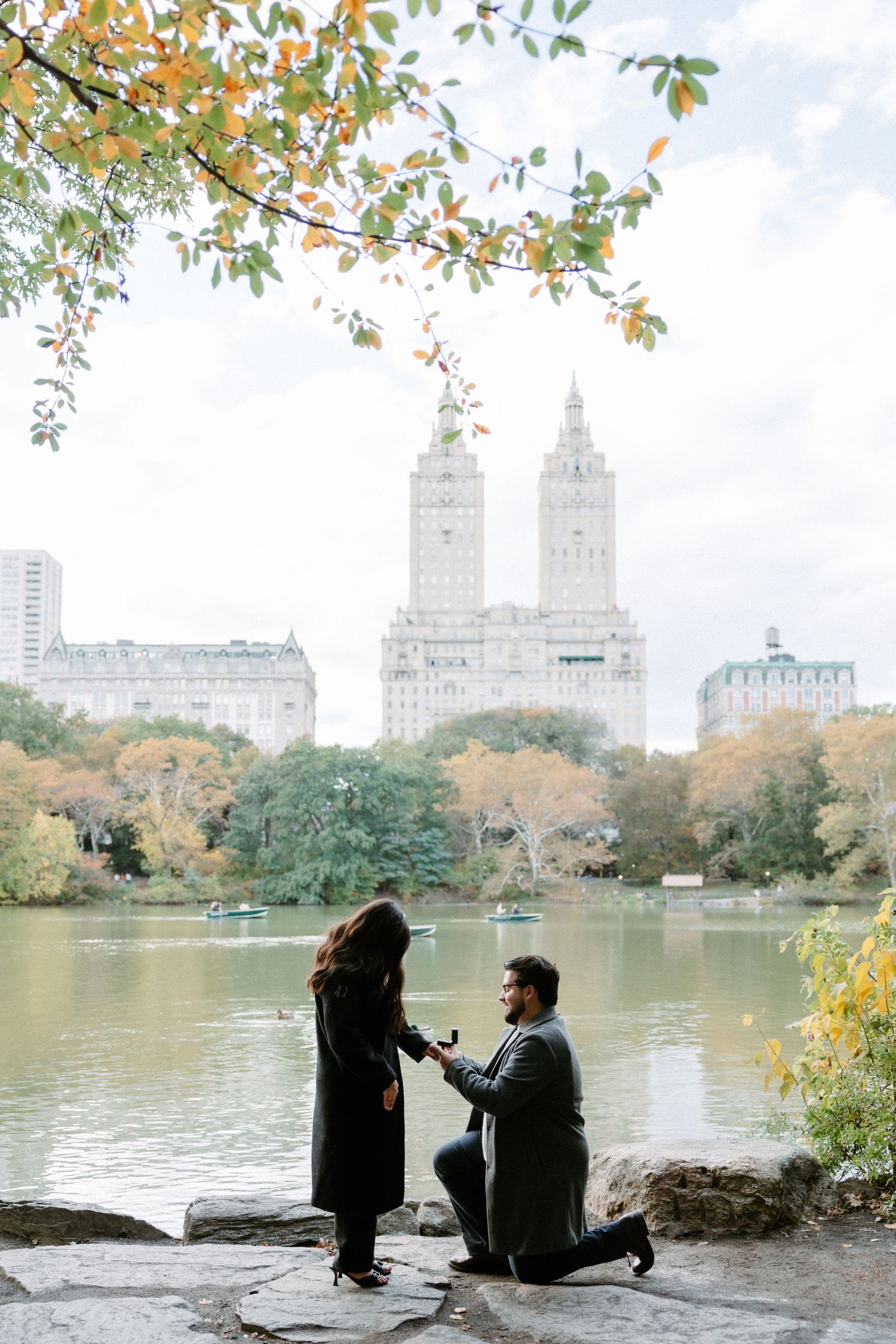 Man proposes to woman on one knee by a lake in Central Park, NYC, with autumn foliage and city skyline backdrop.