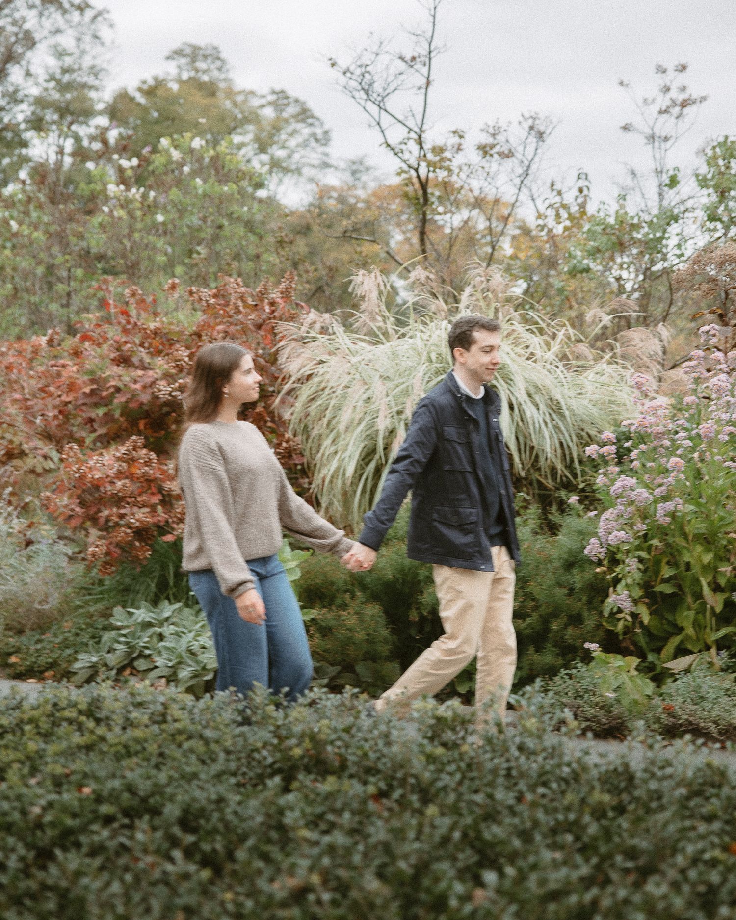 Engaged couple walking hand in hand through a lush garden with autumn foliage.