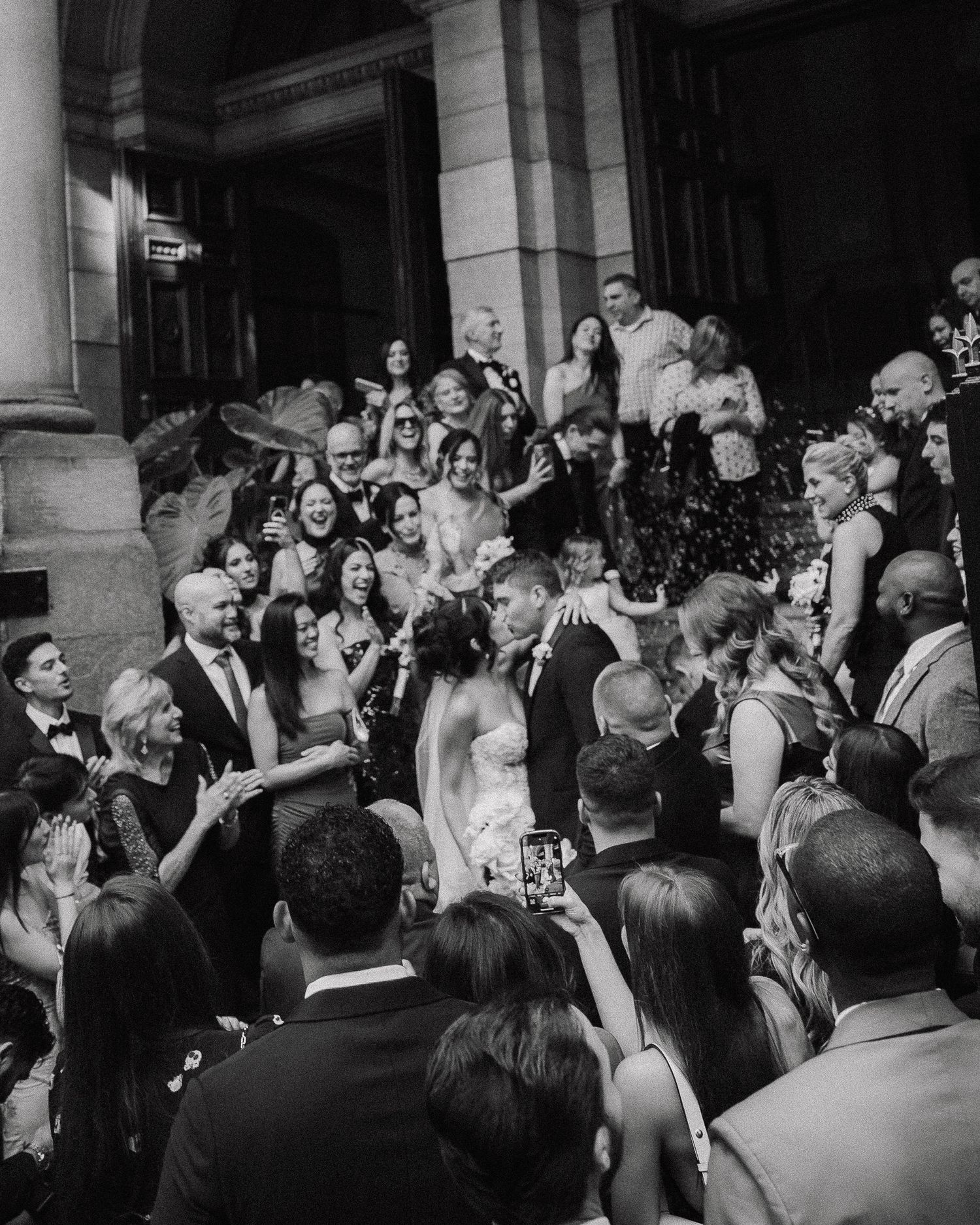 Black and white wedding photo of couple kissing on grand steps surrounded by cheering guests.