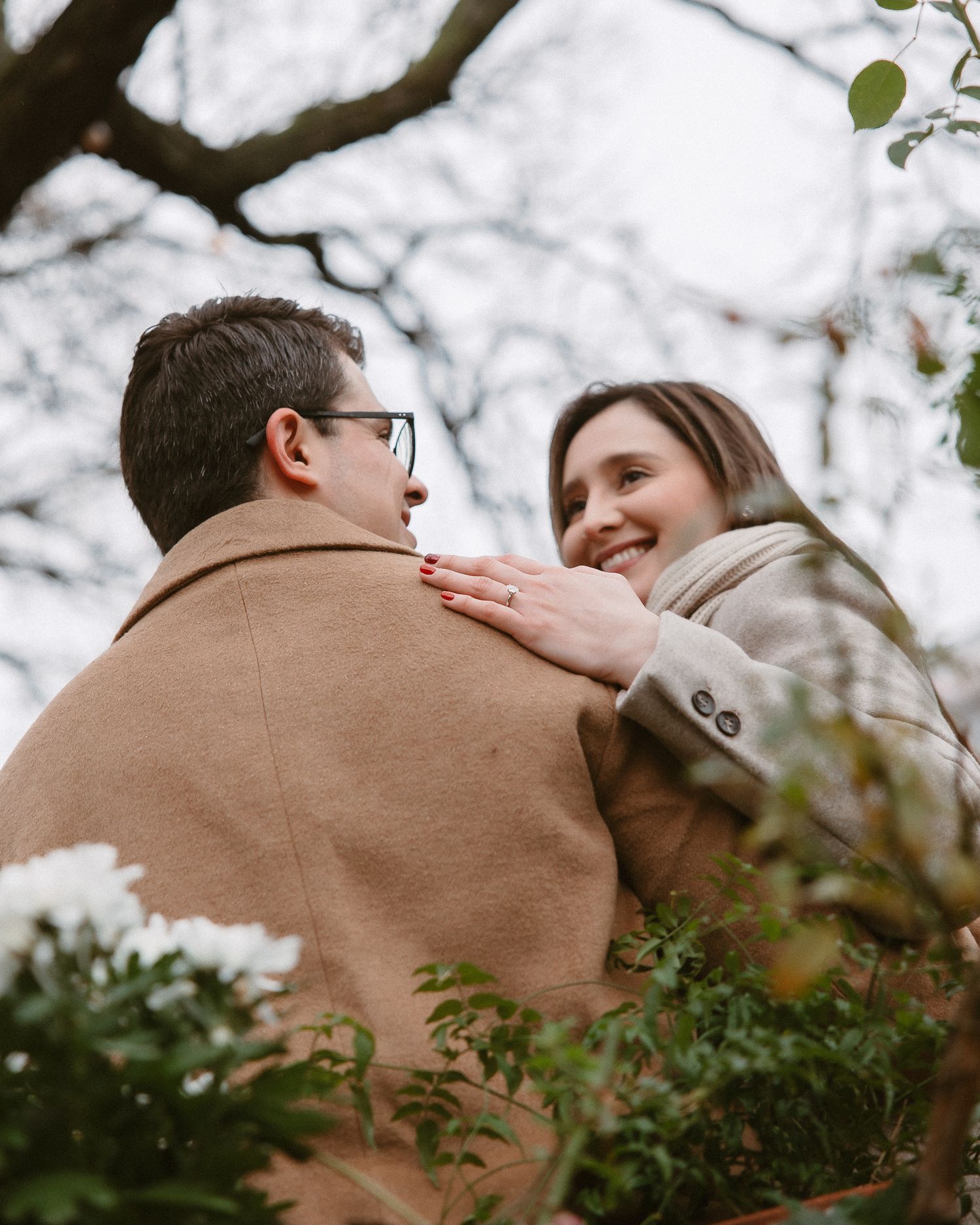 Smiling woman resting hand on partner's shoulder framed by white flowers and bare branches.