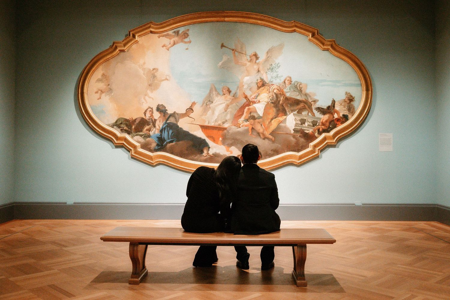 Couple sitting on a bench admiring a large Baroque painting in a museum gallery.