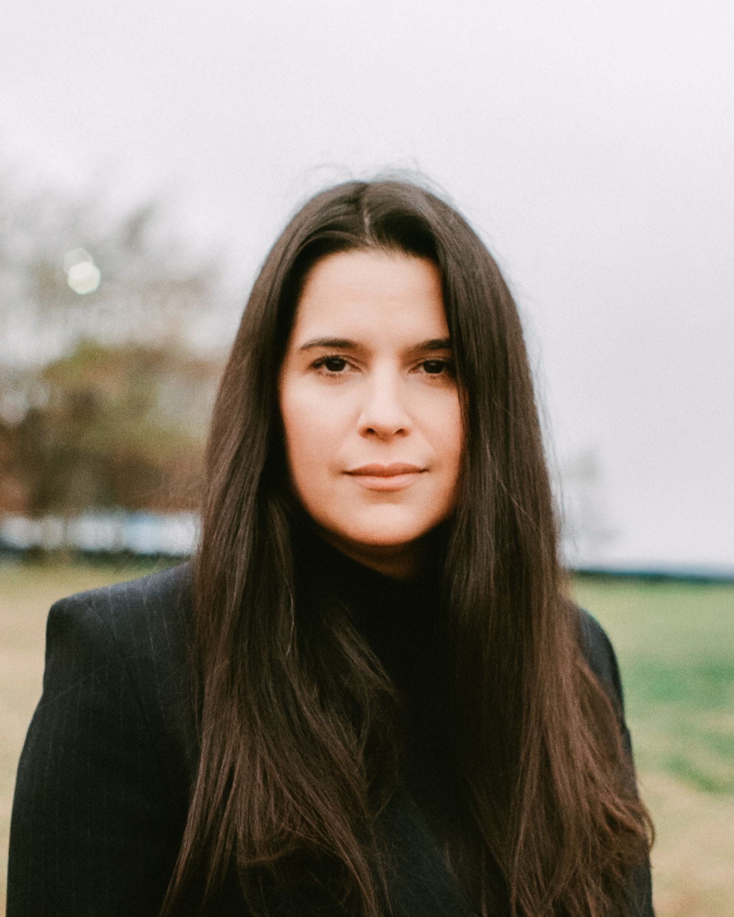 Young woman with long dark hair wearing black turtleneck outdoors with blurred green background.
