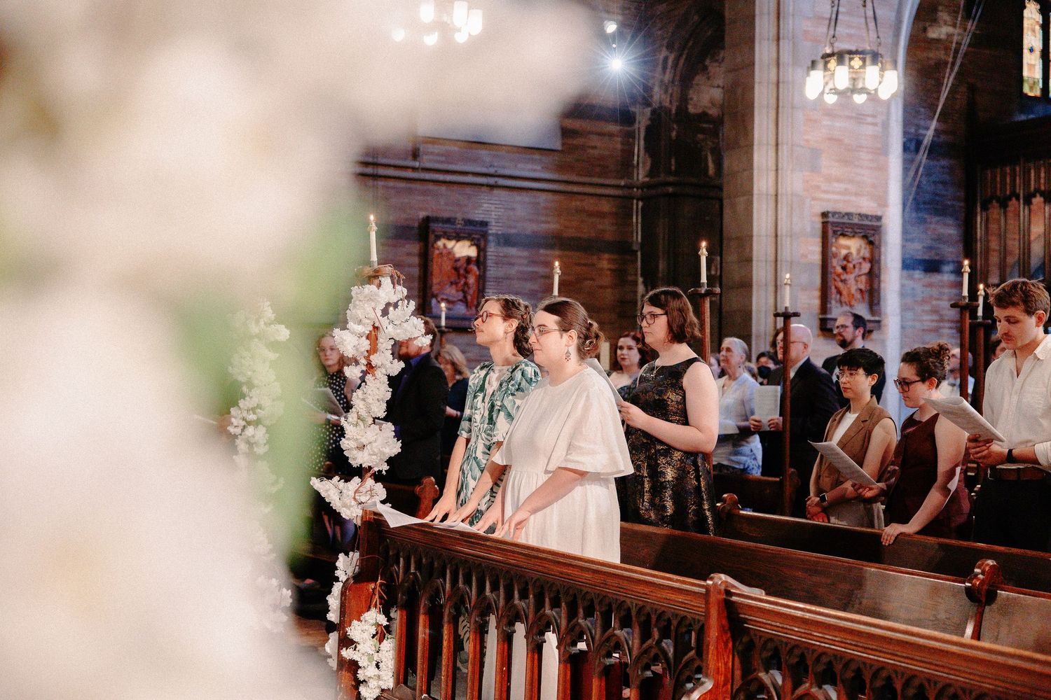 Guests standing in wooden church pews during a wedding ceremony, surrounded by white floral arrangements.