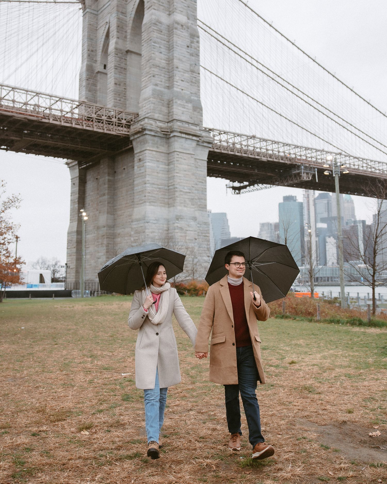 Couple holding umbrellas and hands while walking near Brooklyn Bridge tower on overcast day.