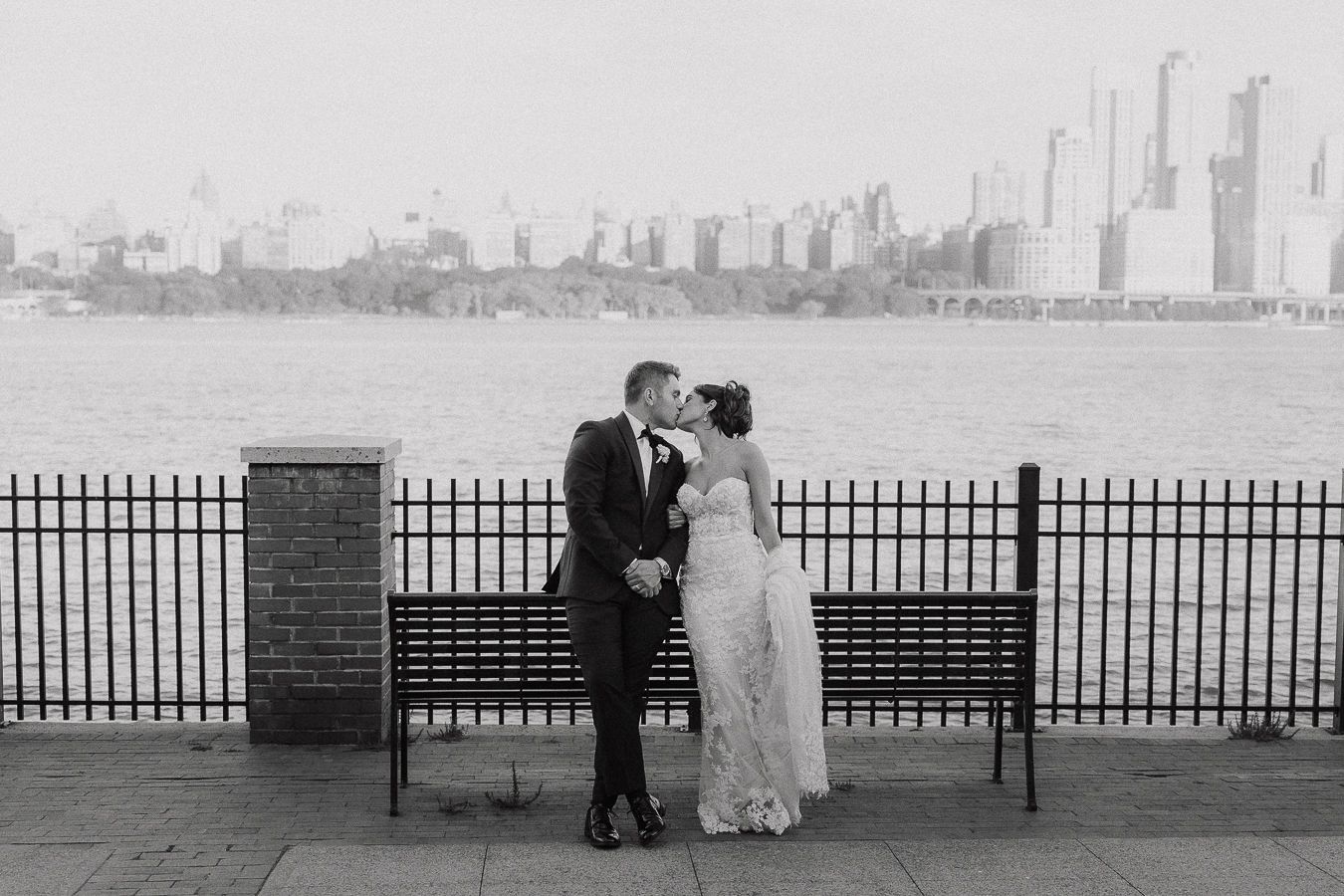 Wedding couple kissing by iron fence with urban skyline and river backdrop, elegant black and white photo.
