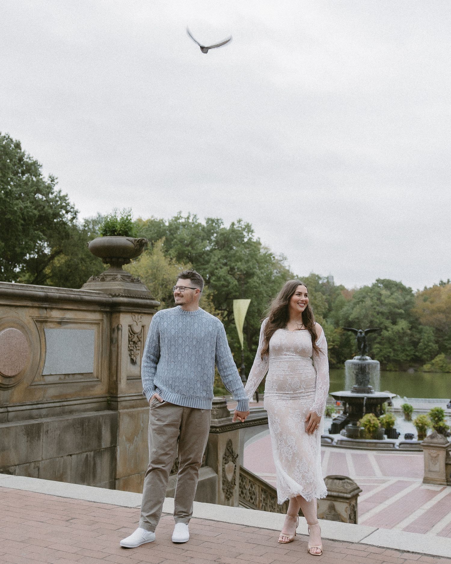 Engaged couple holding hands walking near Bethesda Fountain in Central Park with a white bird flying overhead.