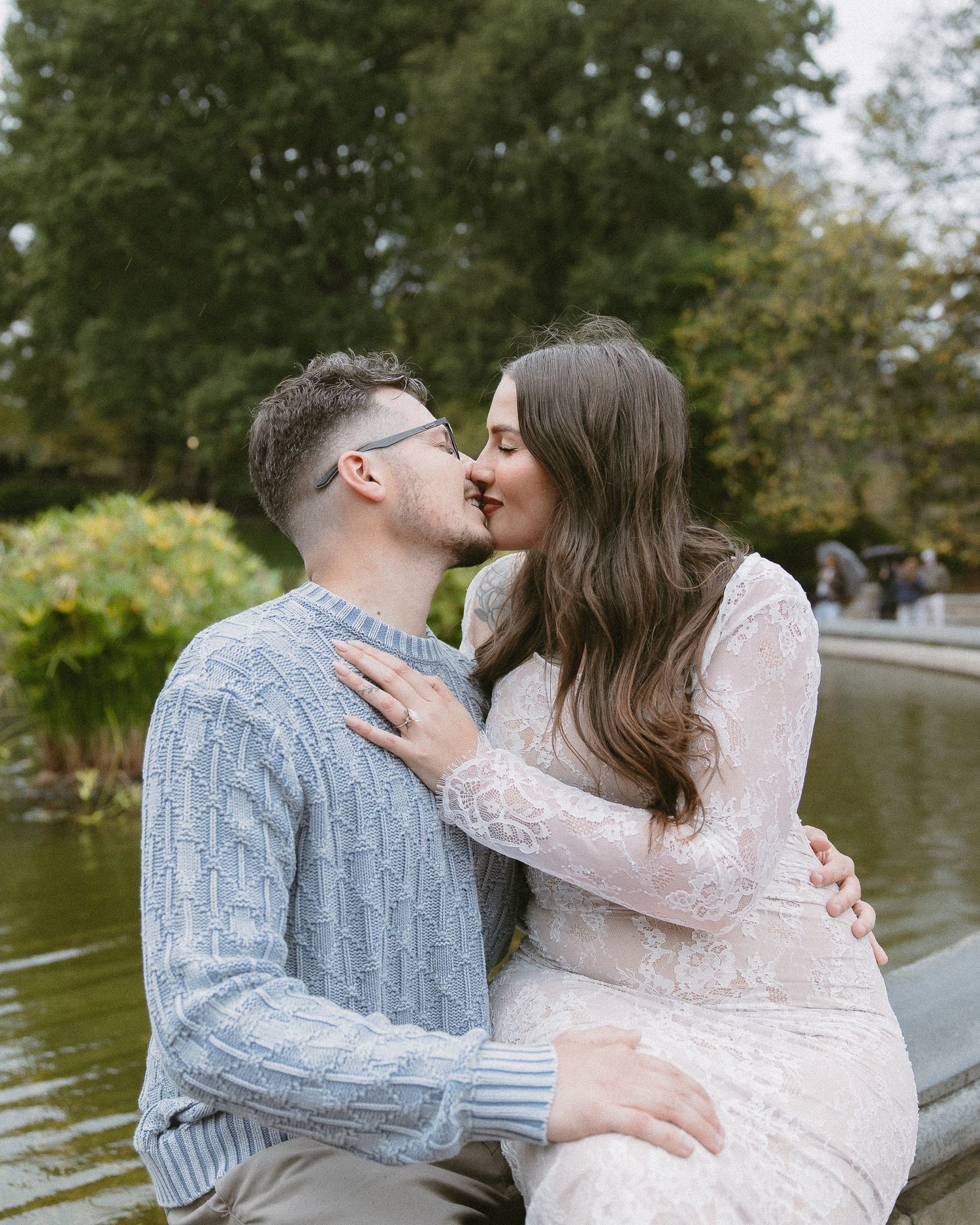 Couple sharing a romantic kiss near a pond in Central Park, woman wearing diamond engagement ring, man in blue sweater.