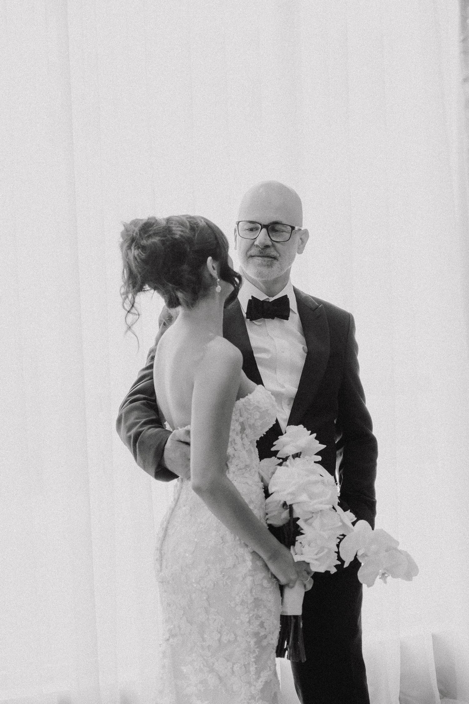 Black and white wedding photo of bride holding bouquet facing groom in tuxedo and bow tie near draped curtains.