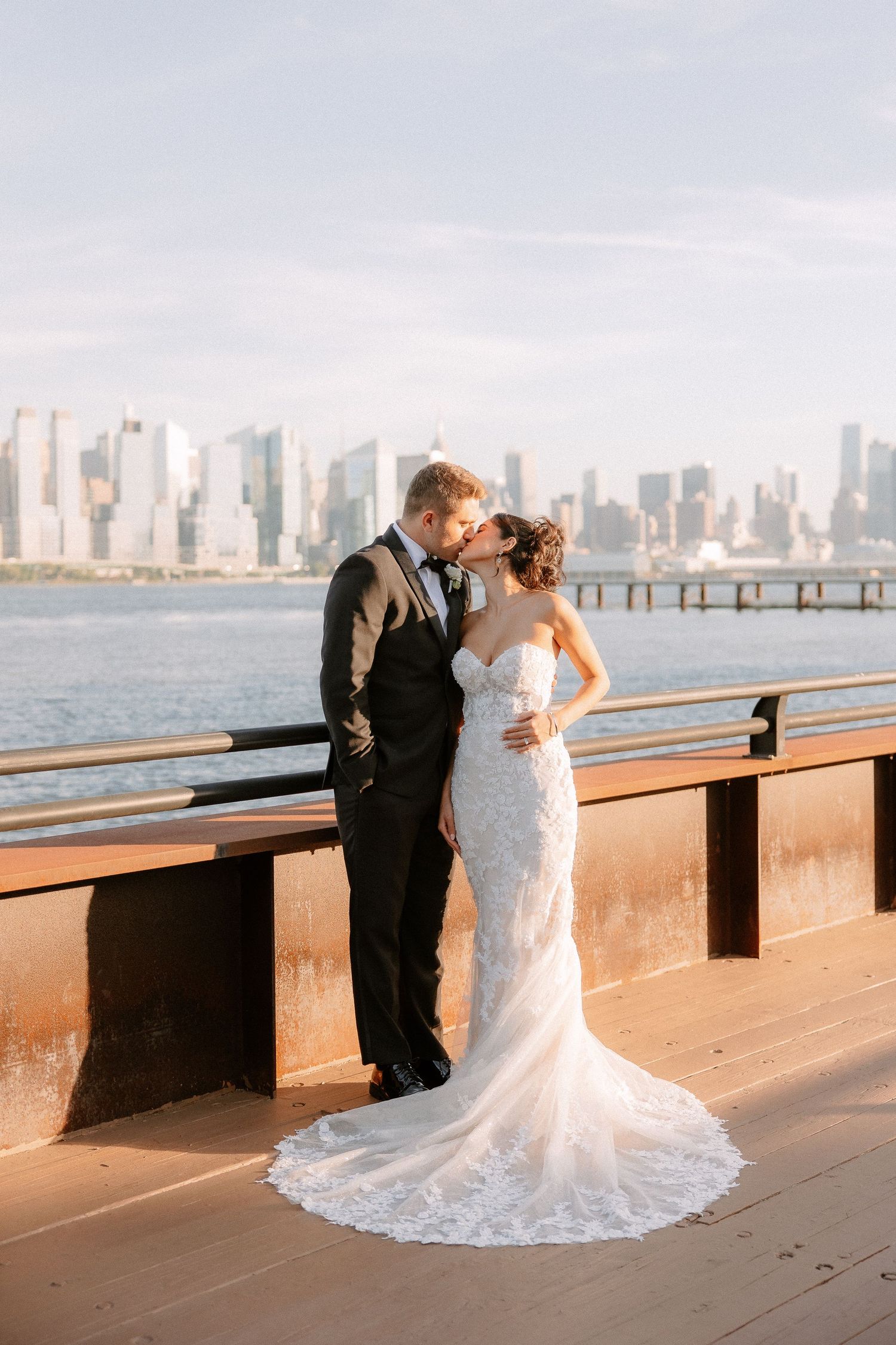 Bride and groom share a kiss on a waterfront deck with the New York City skyline backdrop at golden hour.