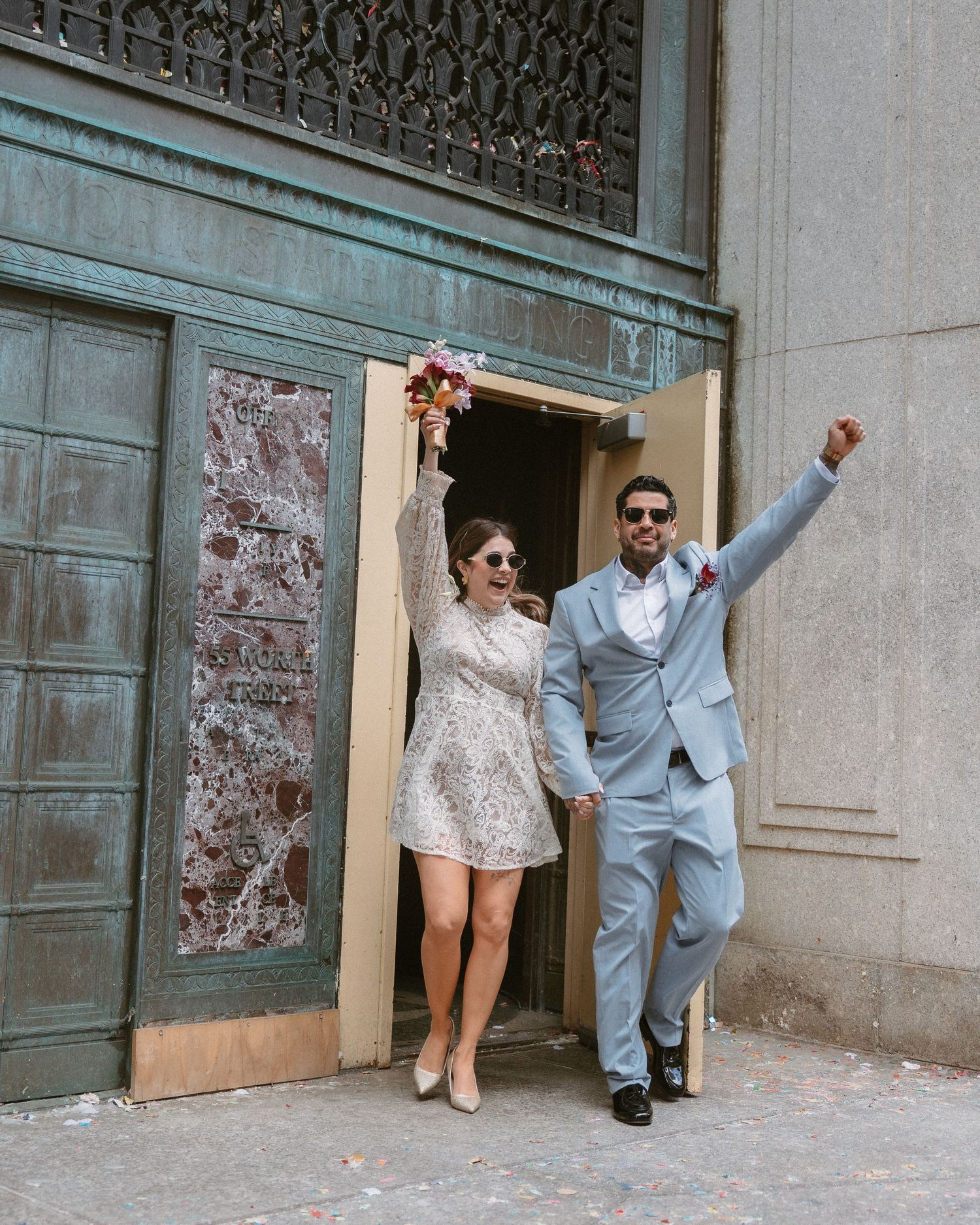 Joyful newlyweds celebrating outside a building, bride in lace mini dress holding bouquet, groom in light blue suit.