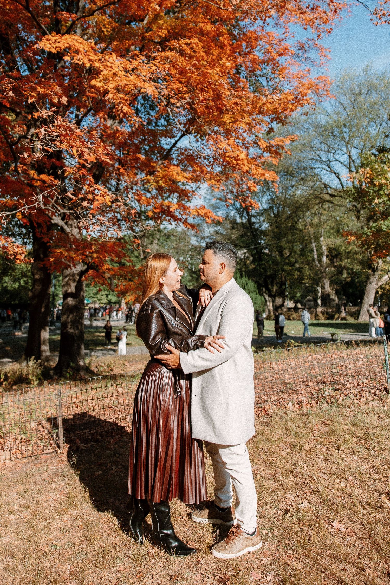 Couple embracing in autumn park with vibrant orange foliage, woman in brown dress, man in white suit.