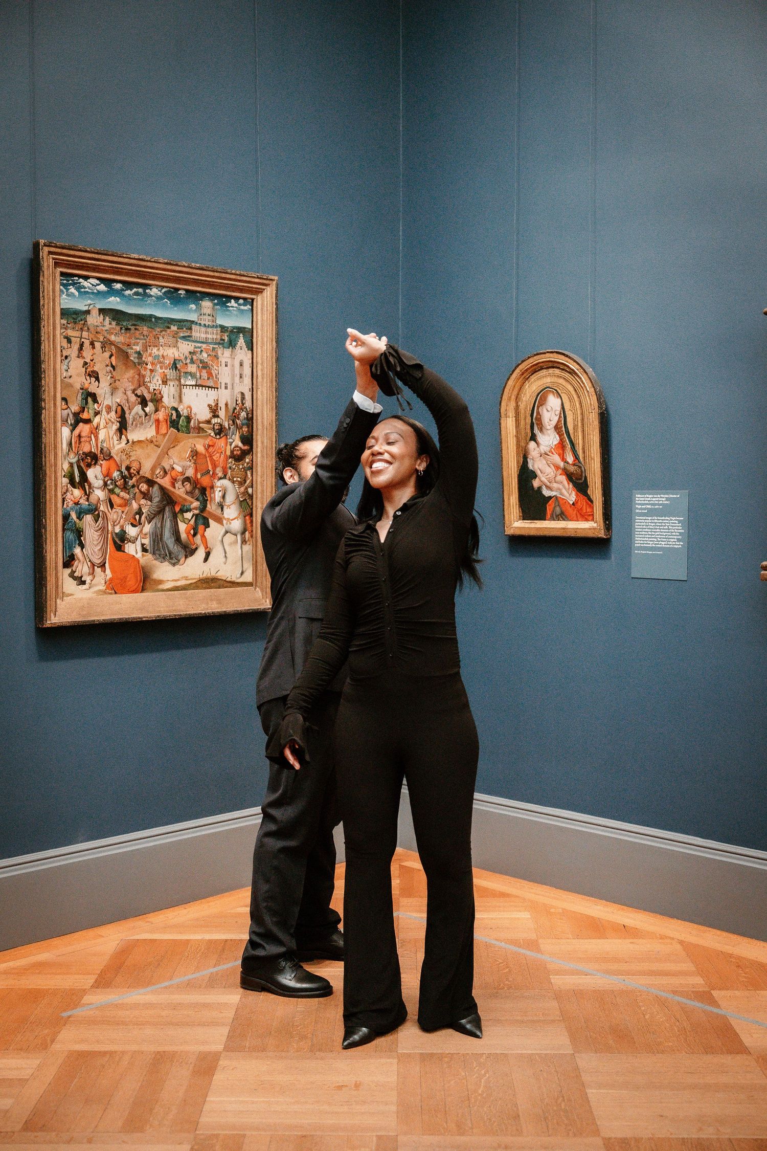 Couple dancing in black outfits inside an art museum gallery with classical paintings on blue walls.