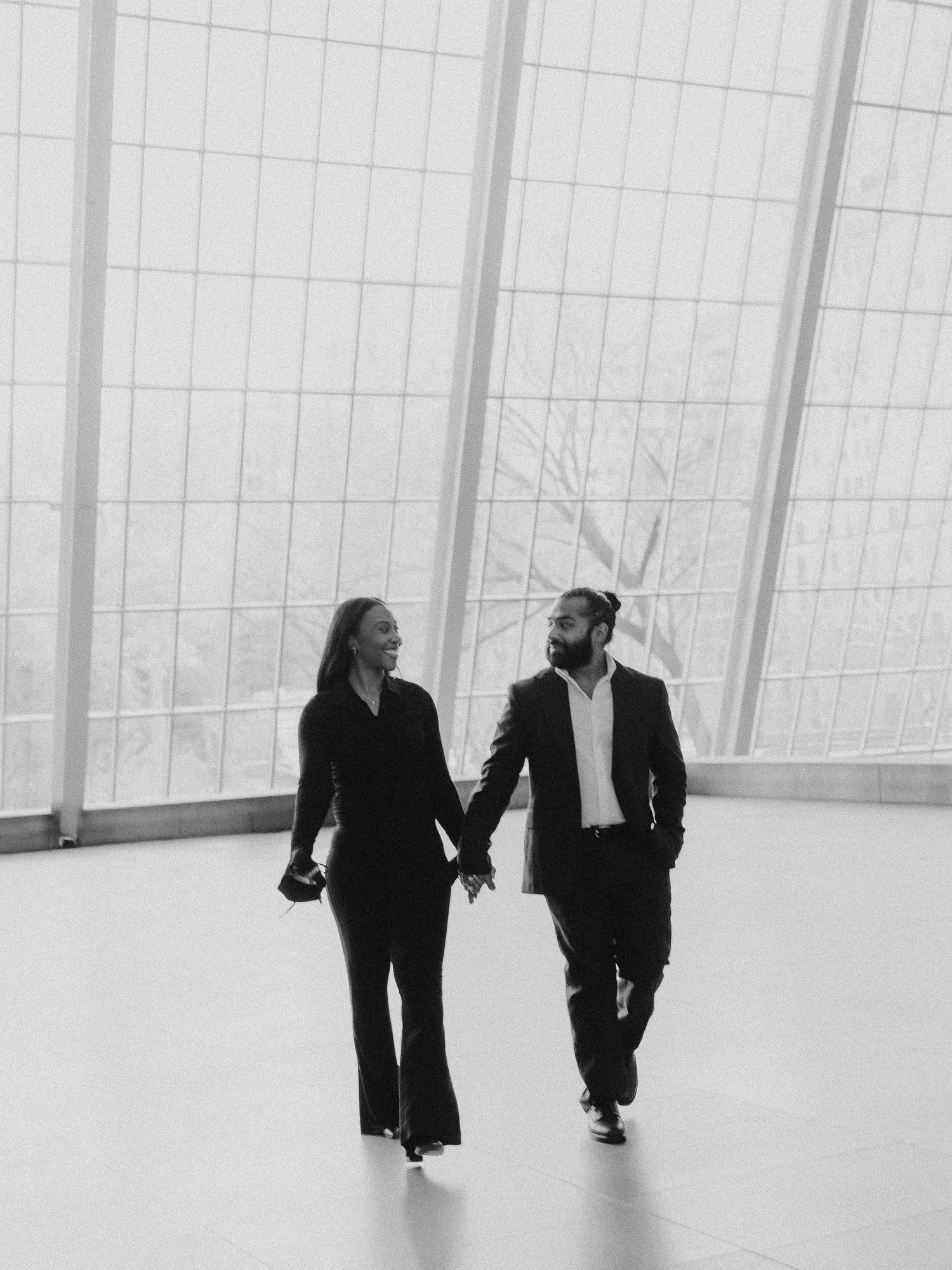 Black and white photo of a couple holding hands and walking together in a modern building with large windows.