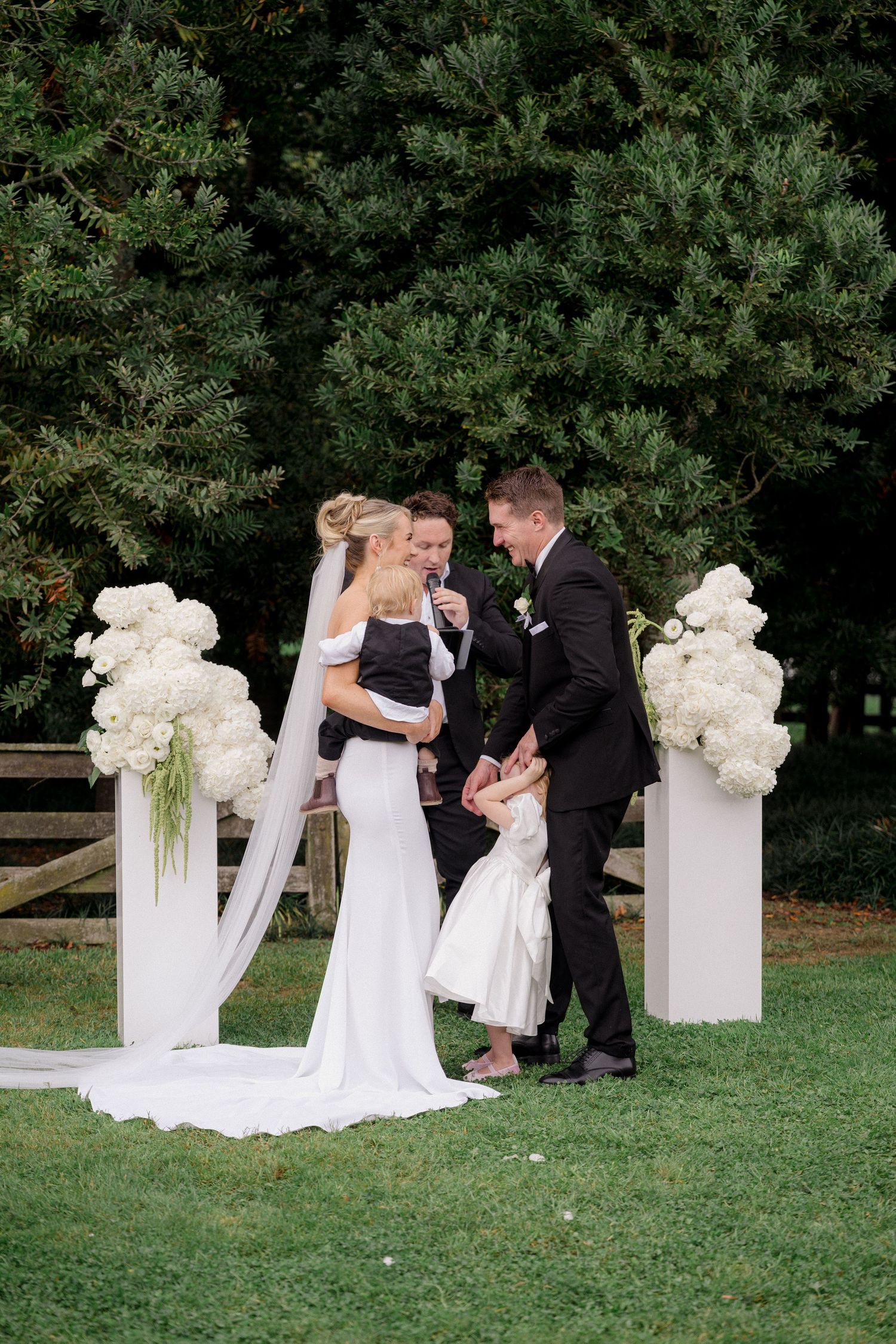 Bride and groom exchange vows outdoors with children present at white floral altar.