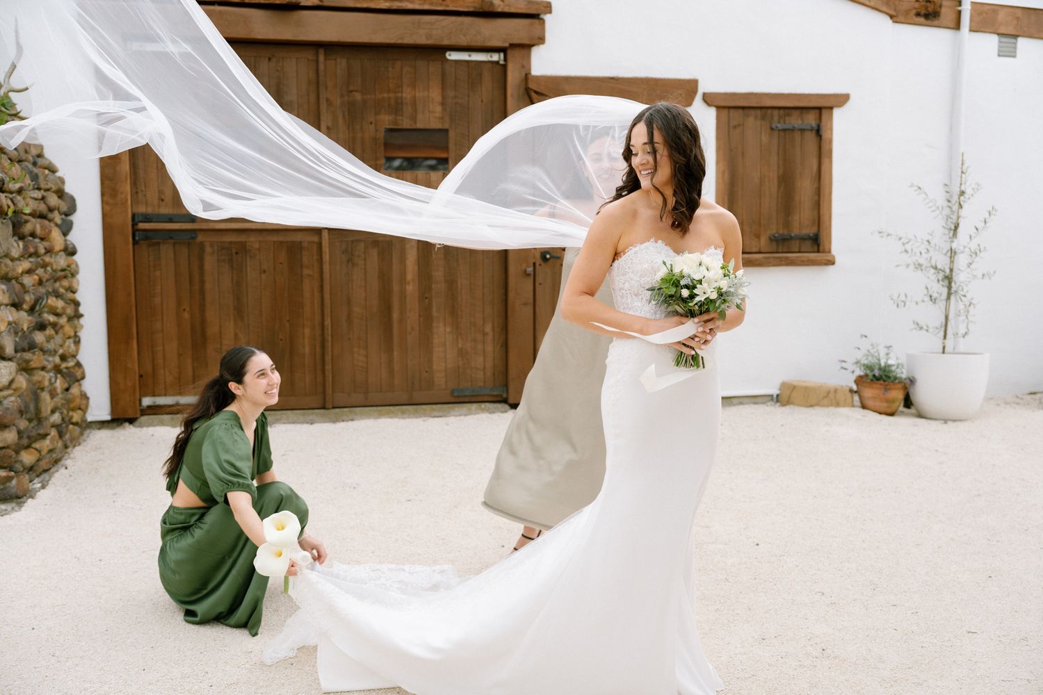 Bridesmaid in green dress adjusts brides flowing veil outside rustic wooden barn as bride holds white bouquet.