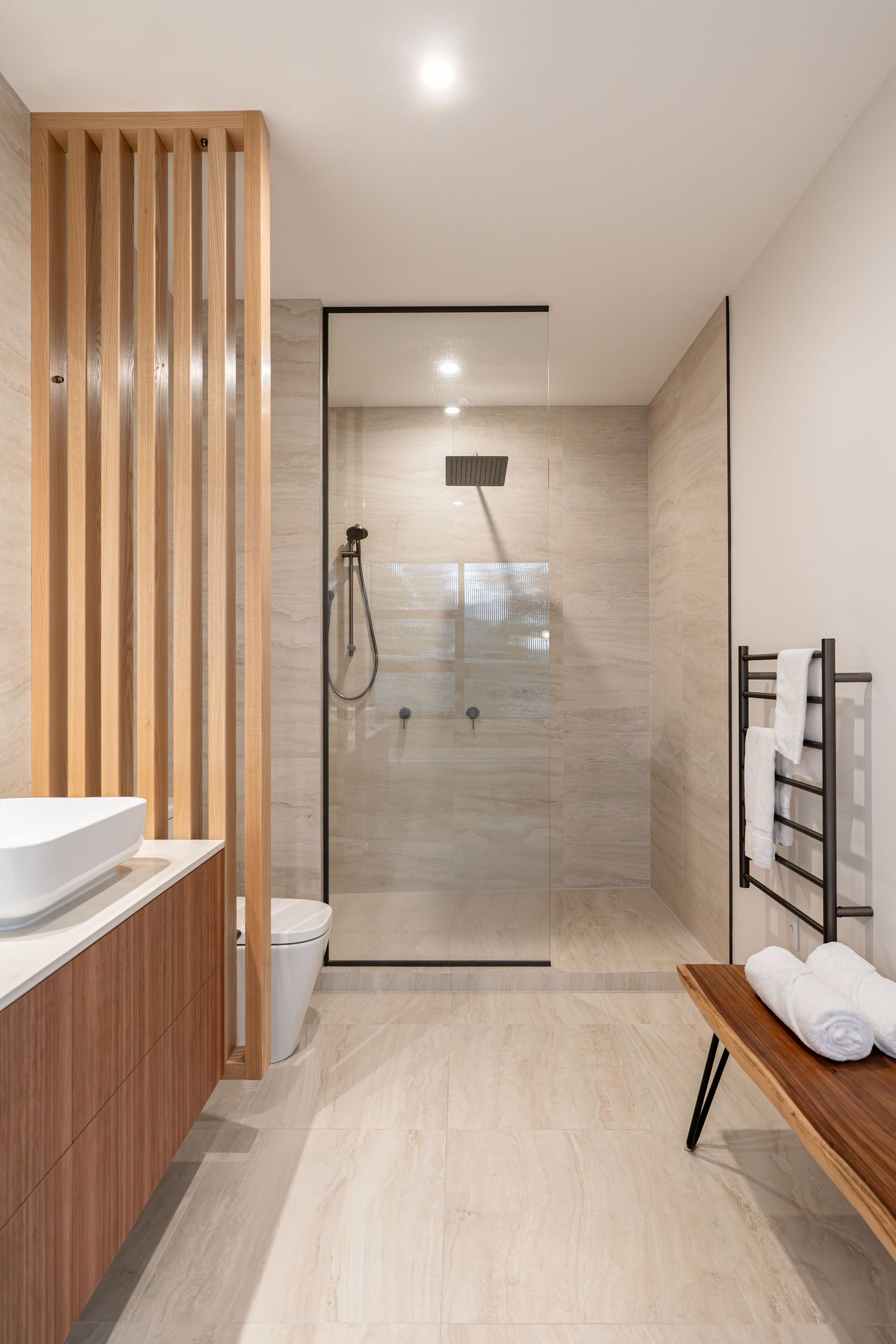 Modern bathroom with walk-in glass shower, wooden vanity, heated towel rail, and neutral stone tiles.
