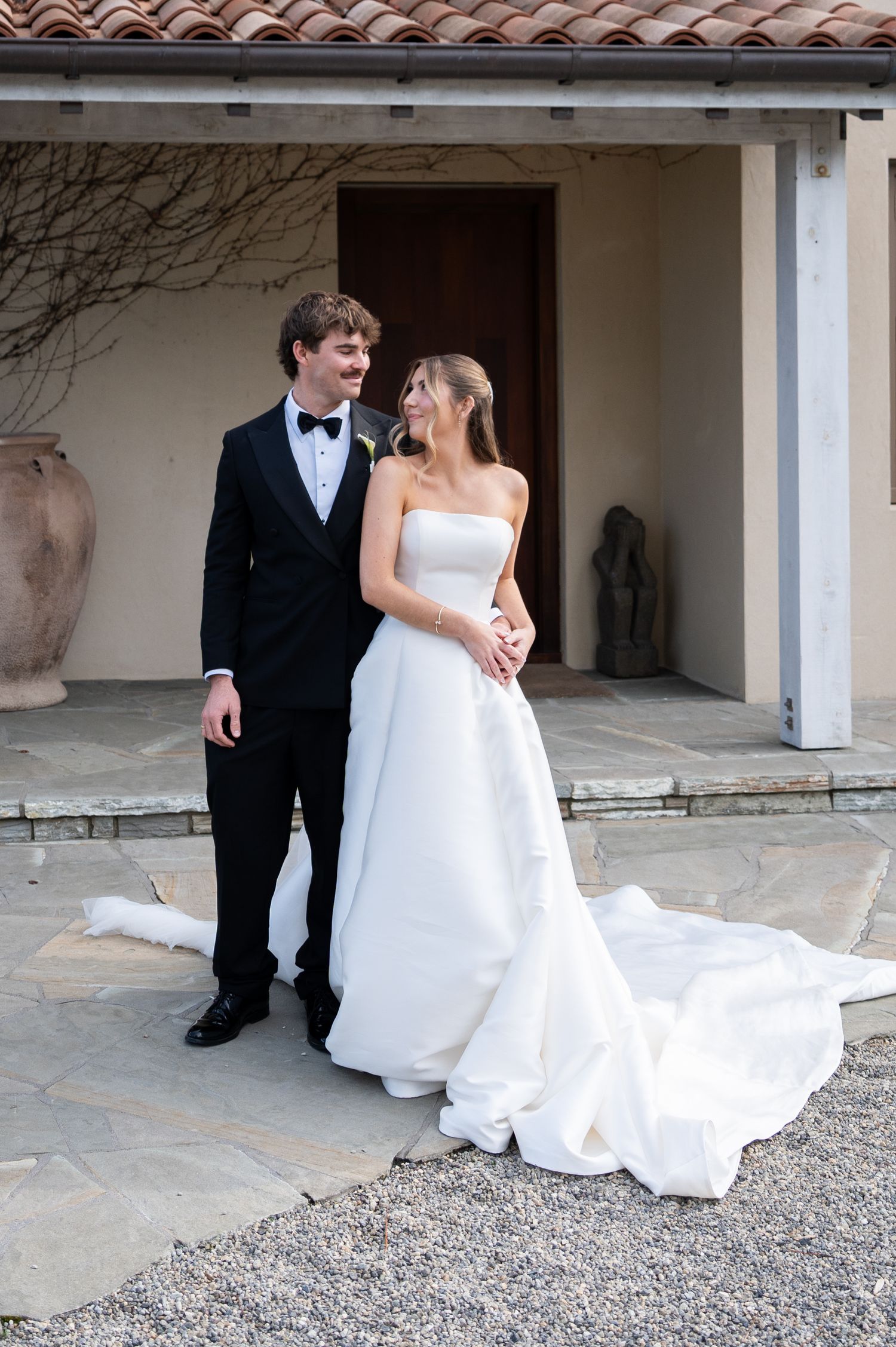 Bride in strapless white gown with train stands with groom in black tuxedo outside elegant villa.