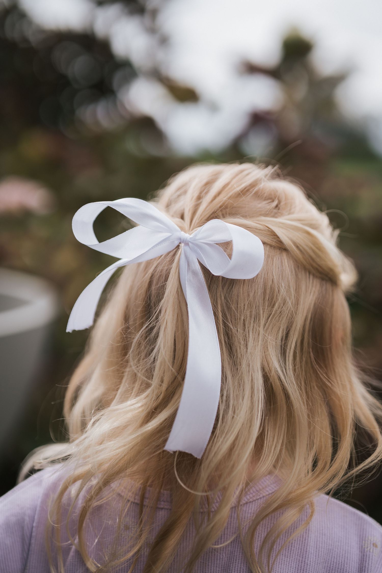 Blonde girl with elegant white ribbon bow hairstyle viewed from behind in garden setting.