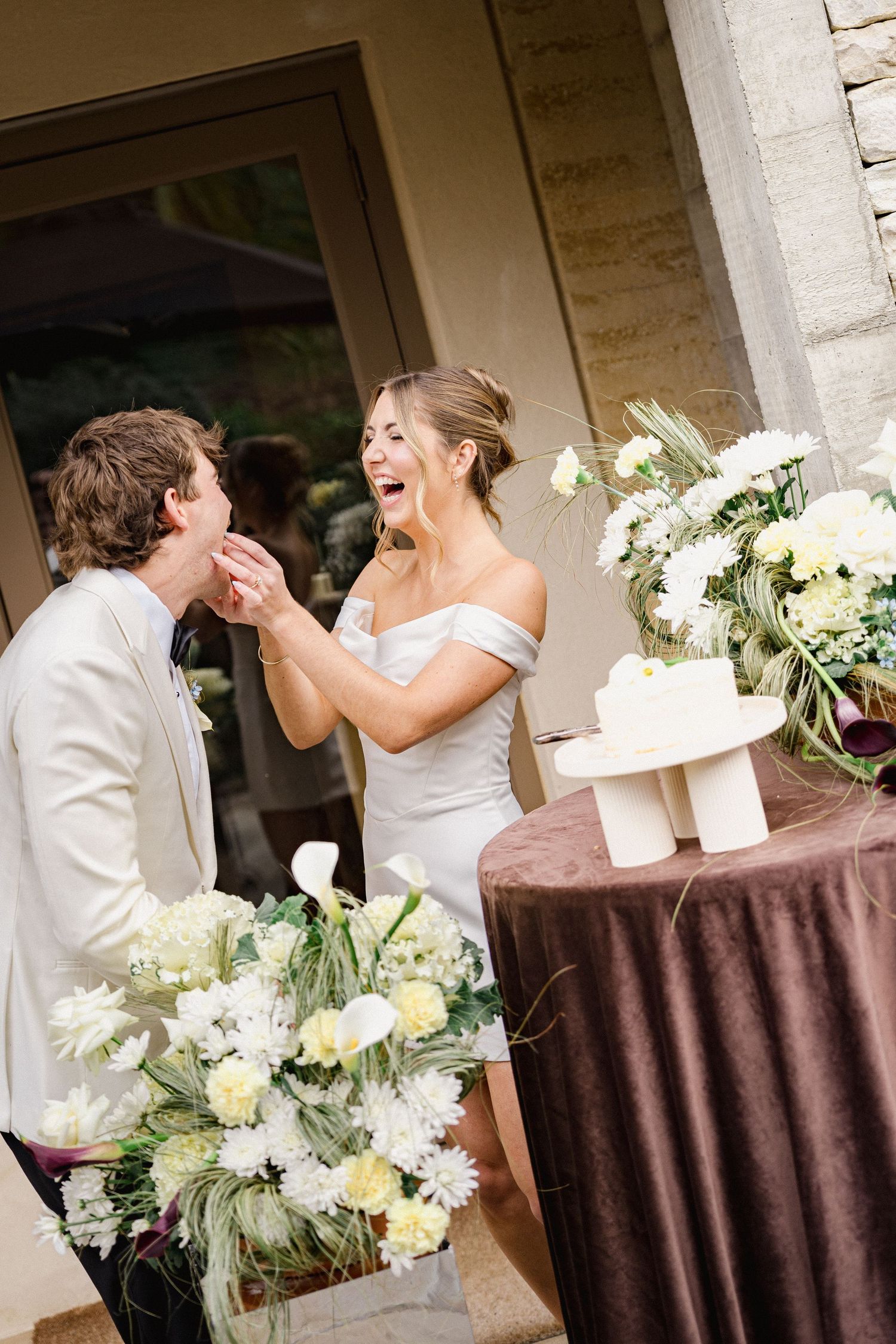 Laughing bride in off-shoulder gown feeds cake to groom beside white floral arrangements at reception.