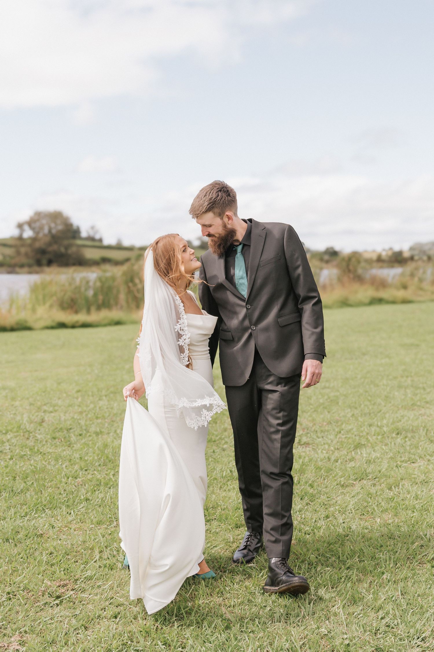 Bride in white dress with lace veil and bearded groom in gray suit share intimate moment outdoors.