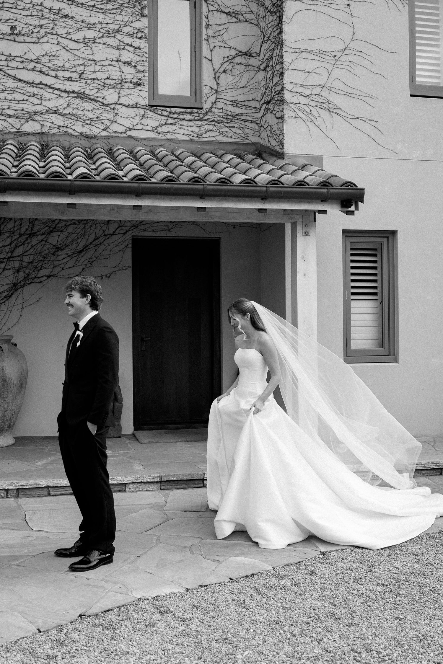 Black and white photo of bride in flowing veil and groom outside a Mediterranean-style wedding venue.