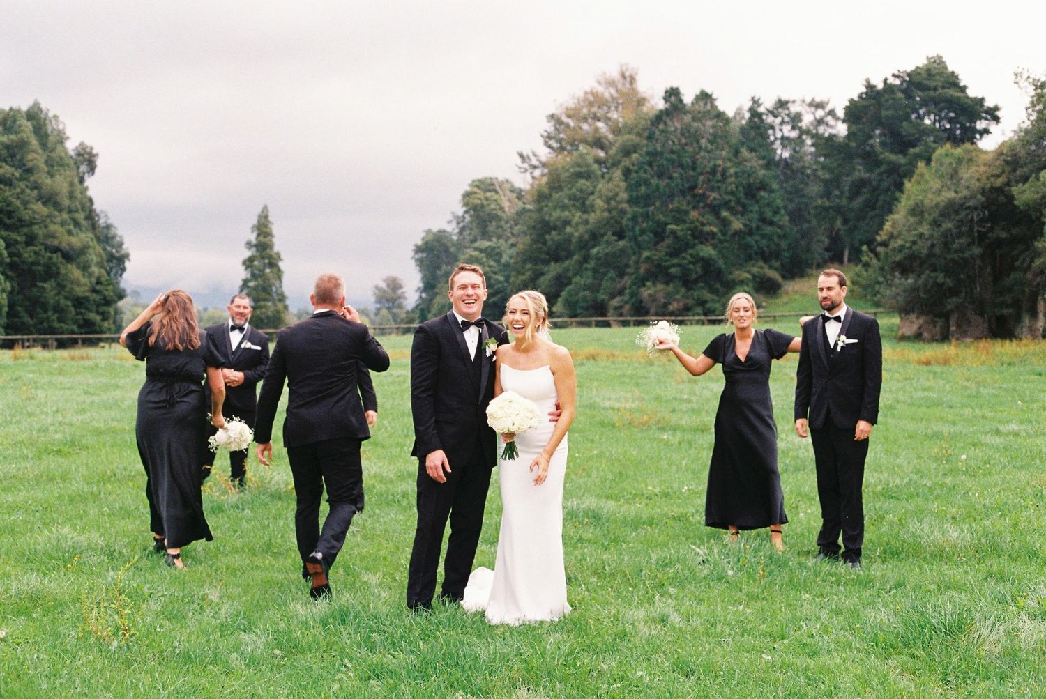 Wedding party in black attire walks across green field with bride and groom at center.
