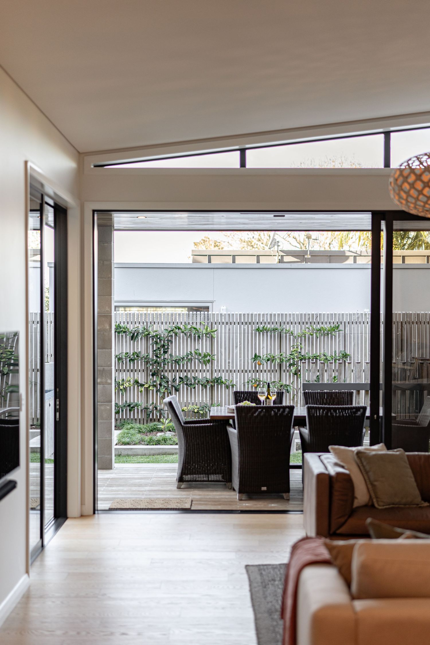 Modern open-plan living room with sliding glass doors opening to an outdoor dining area with rattan chairs and lush garden.