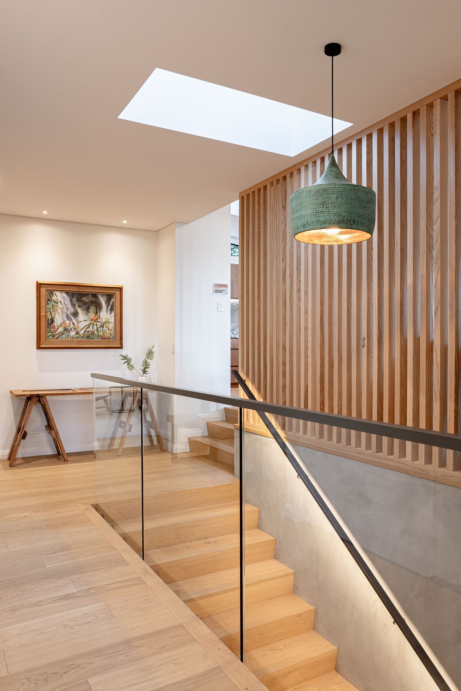 Modern hallway with wooden staircase, glass railing, skylight, pendant lamp, and framed artwork on white wall.