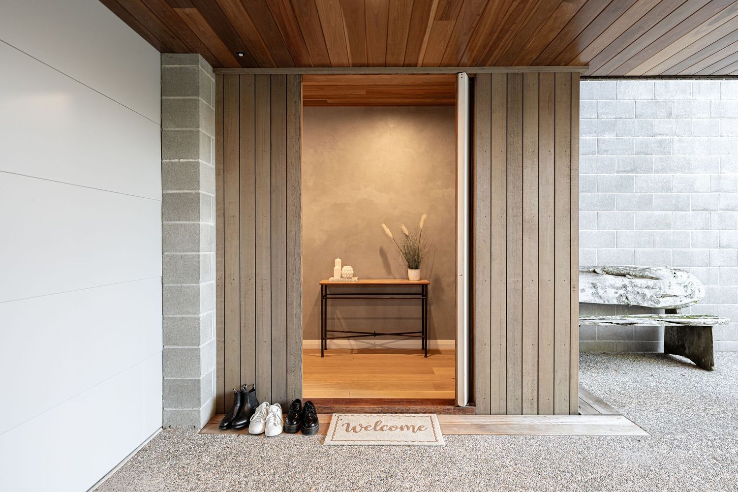 Modern home entryway with wood-paneled walls, welcome mat, shoes, and a console table visible through open door.