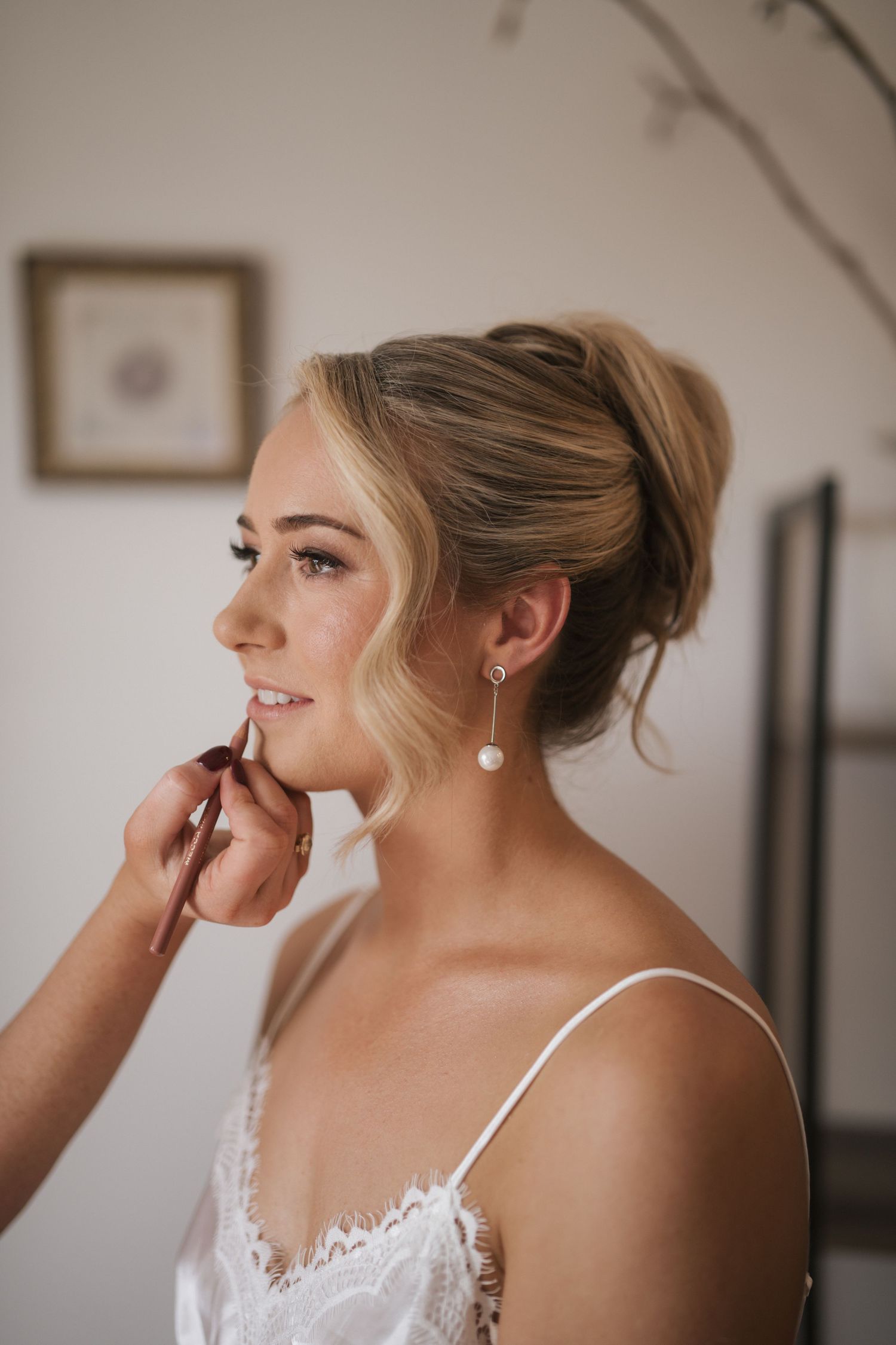 Blonde bride with updo and pearl earrings gets makeup applied during wedding preparations.