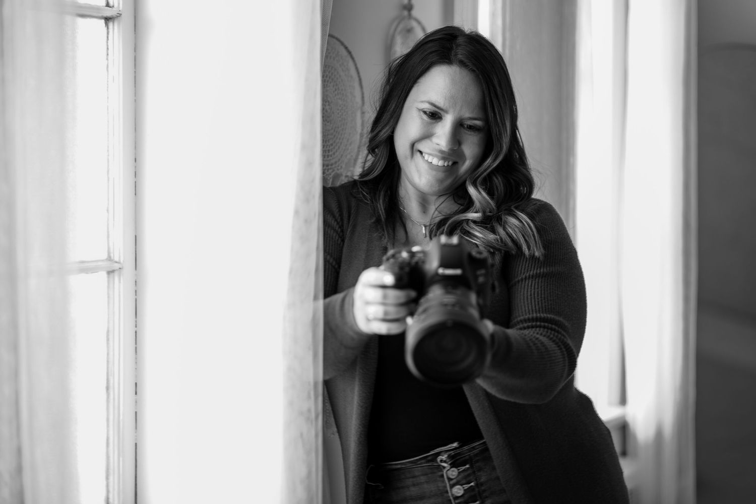 Black and white photo of a smiling woman holding a DSLR camera near a bright window.