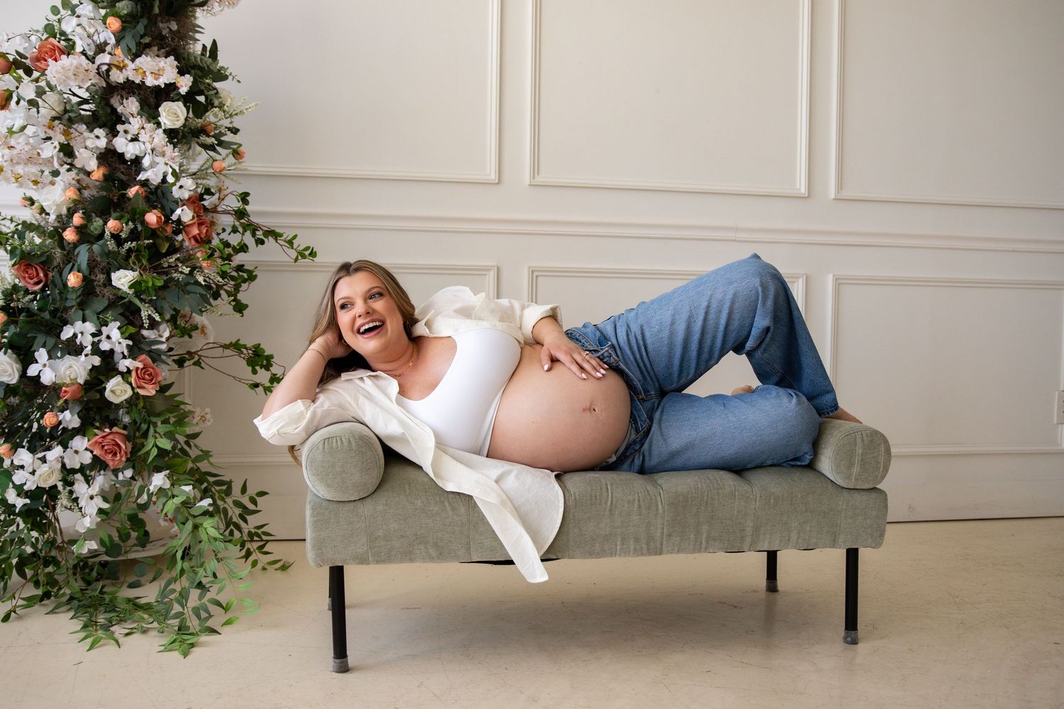 Maternity photo shoot on grey couch with floral arrangements and elegant white decor.