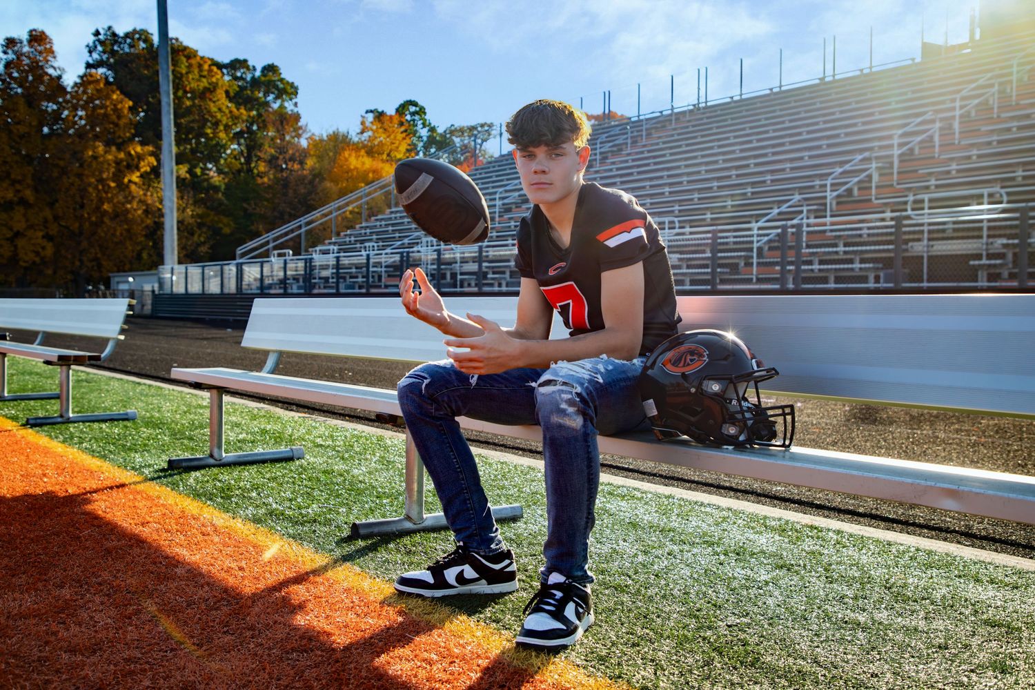 A football player sits on stadium bleachers holding a helmet at sunset.