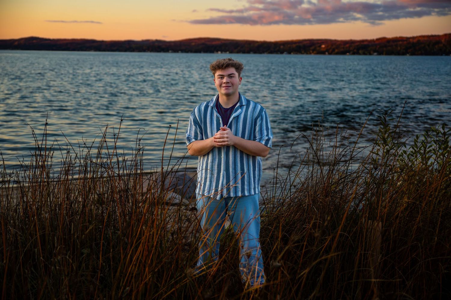 Person in striped shirt poses by tranquil lake at sunset with tall grass in foreground.