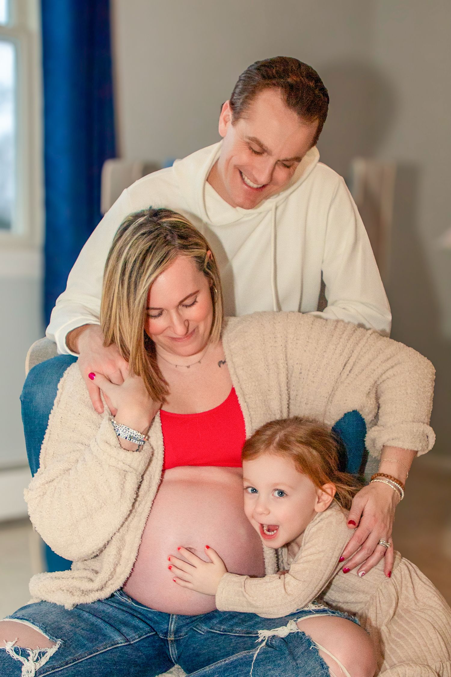 Family maternity photoshoot with a red top and cream cardigan against blue walls.