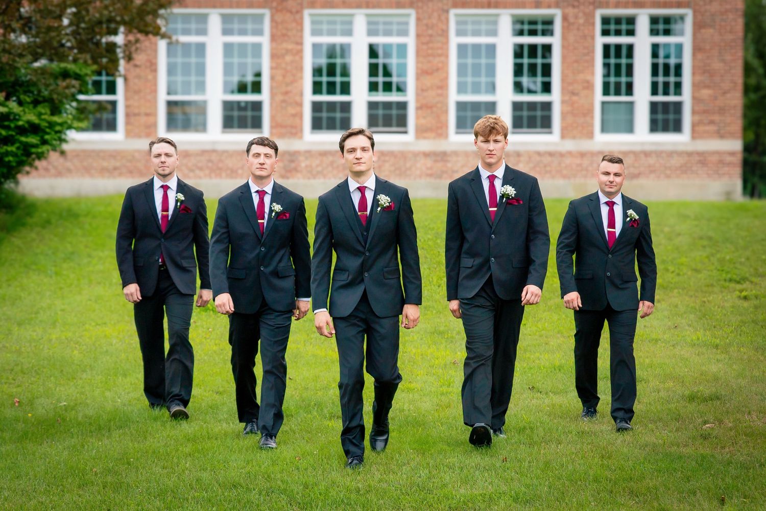Groomsmen in black suits with red ties walking across a green lawn in front of a brick building.