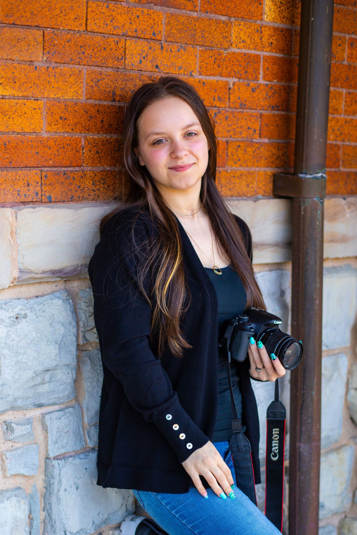 Professional photographer in black blazer holding camera against orange brick and stone wall.