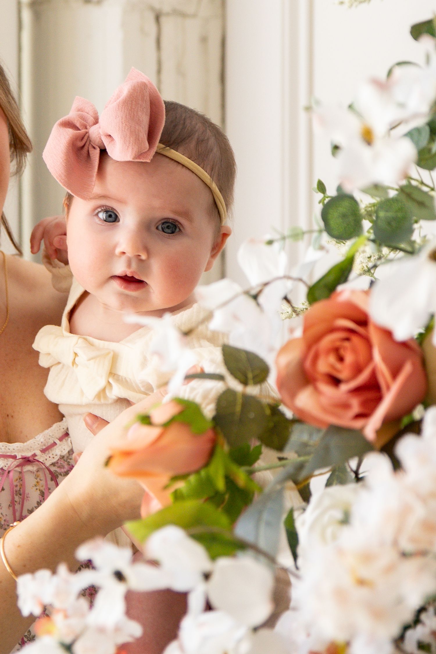 Baby wearing pink bow headband near elegant floral arrangement with peach roses and white flowers.