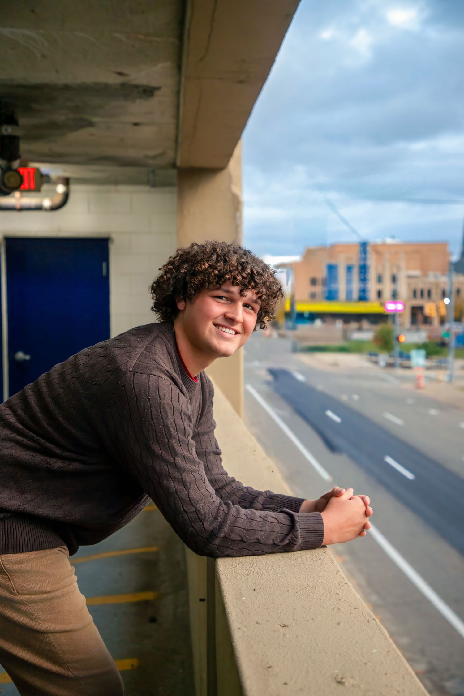 Person leaning against concrete barrier in parking structure with city view.