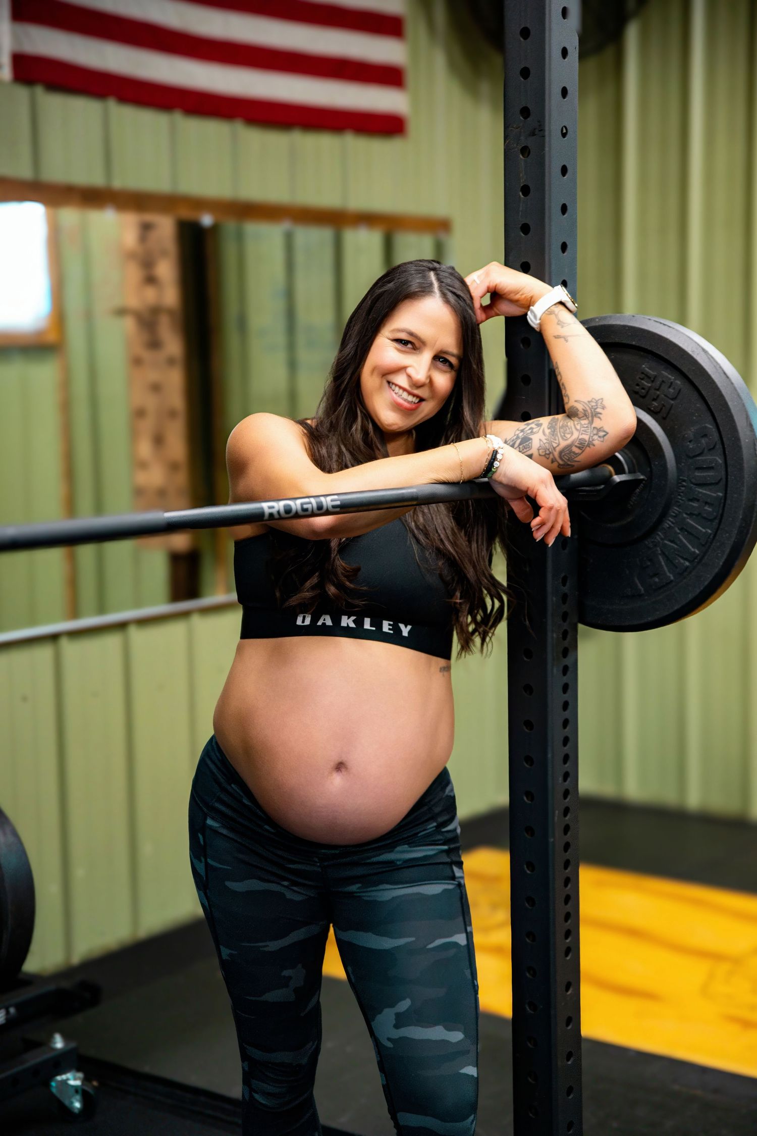 Pregnant person in workout attire poses with barbell in gym with American flag.