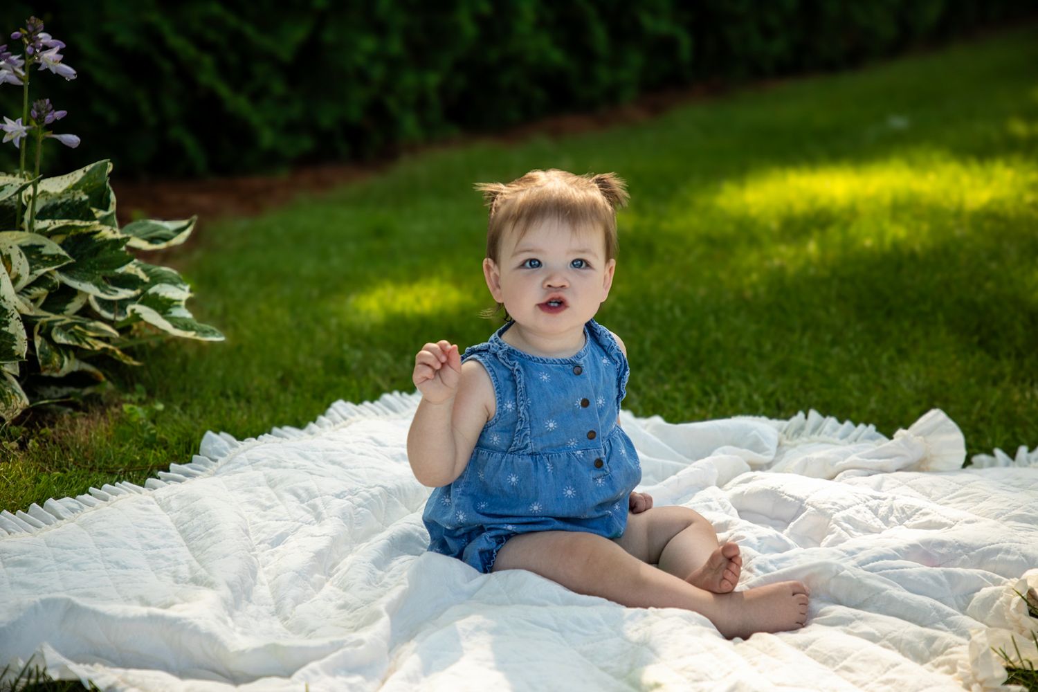 Baby in blue outfit sitting on white blanket in sunny garden setting.