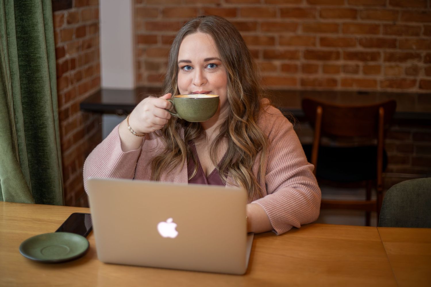 Person in pink sweater drinking from green mug while working on MacBook at cafe.