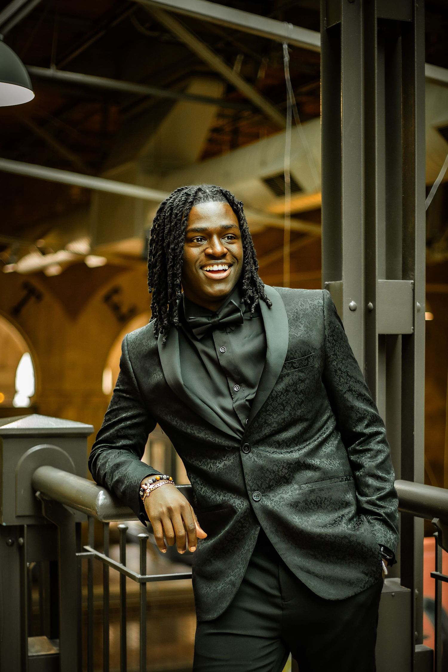 Person in black suit and hoodie smiles while standing in modern industrial setting.