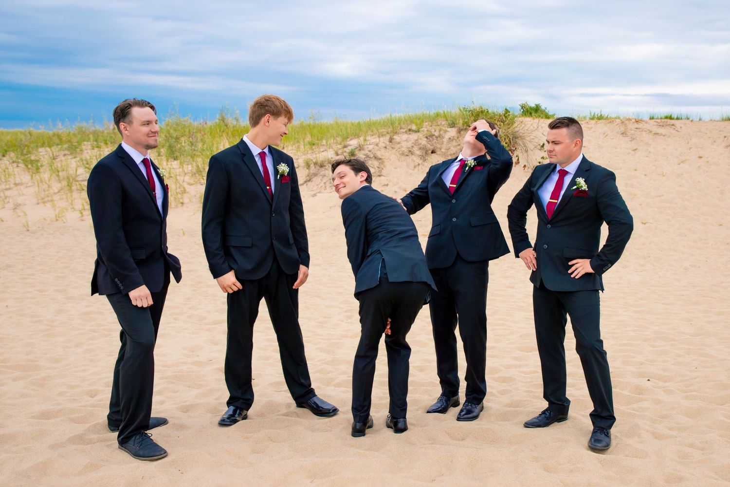 Five groomsmen in dark suits pose playfully on a sandy beach under blue skies.