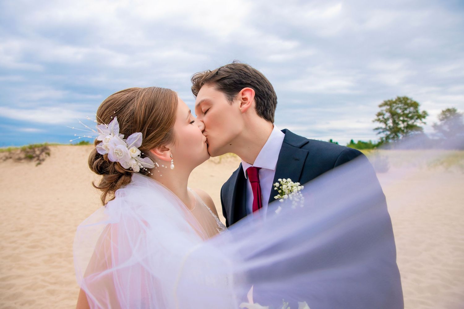 Romantic wedding couple sharing a kiss on a sandy beach under cloudy skies.