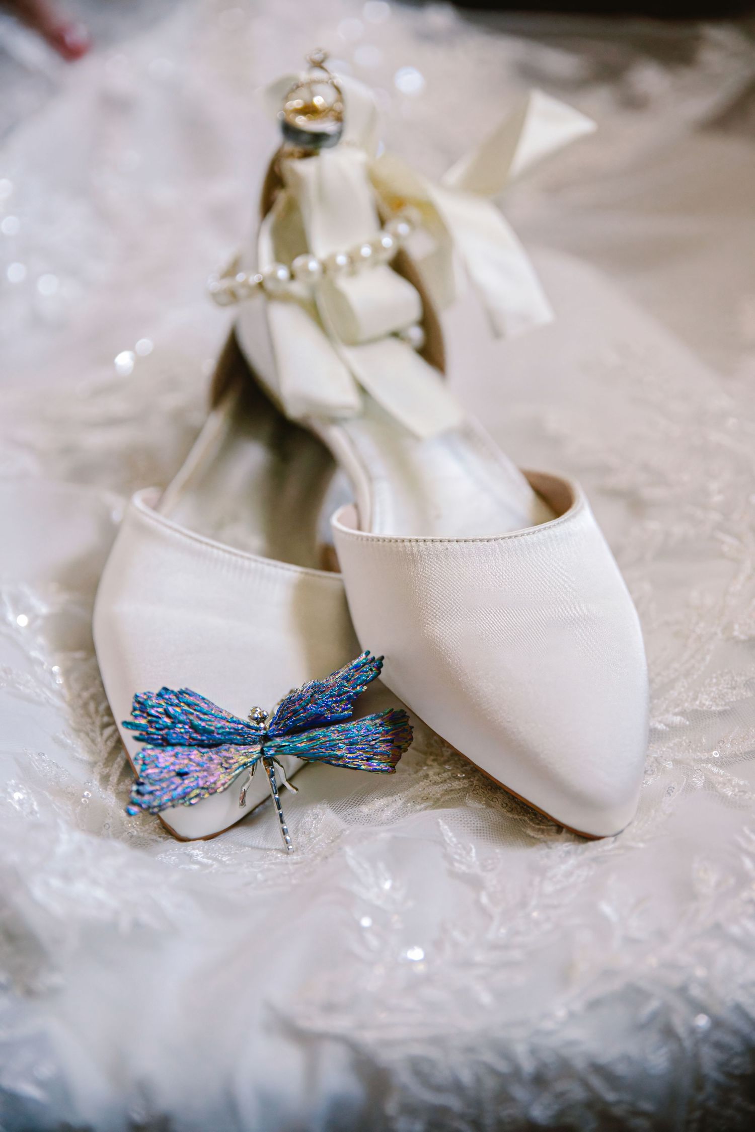Elegant white bridal shoes with a decorative blue dragonfly brooch on delicate lace fabric.