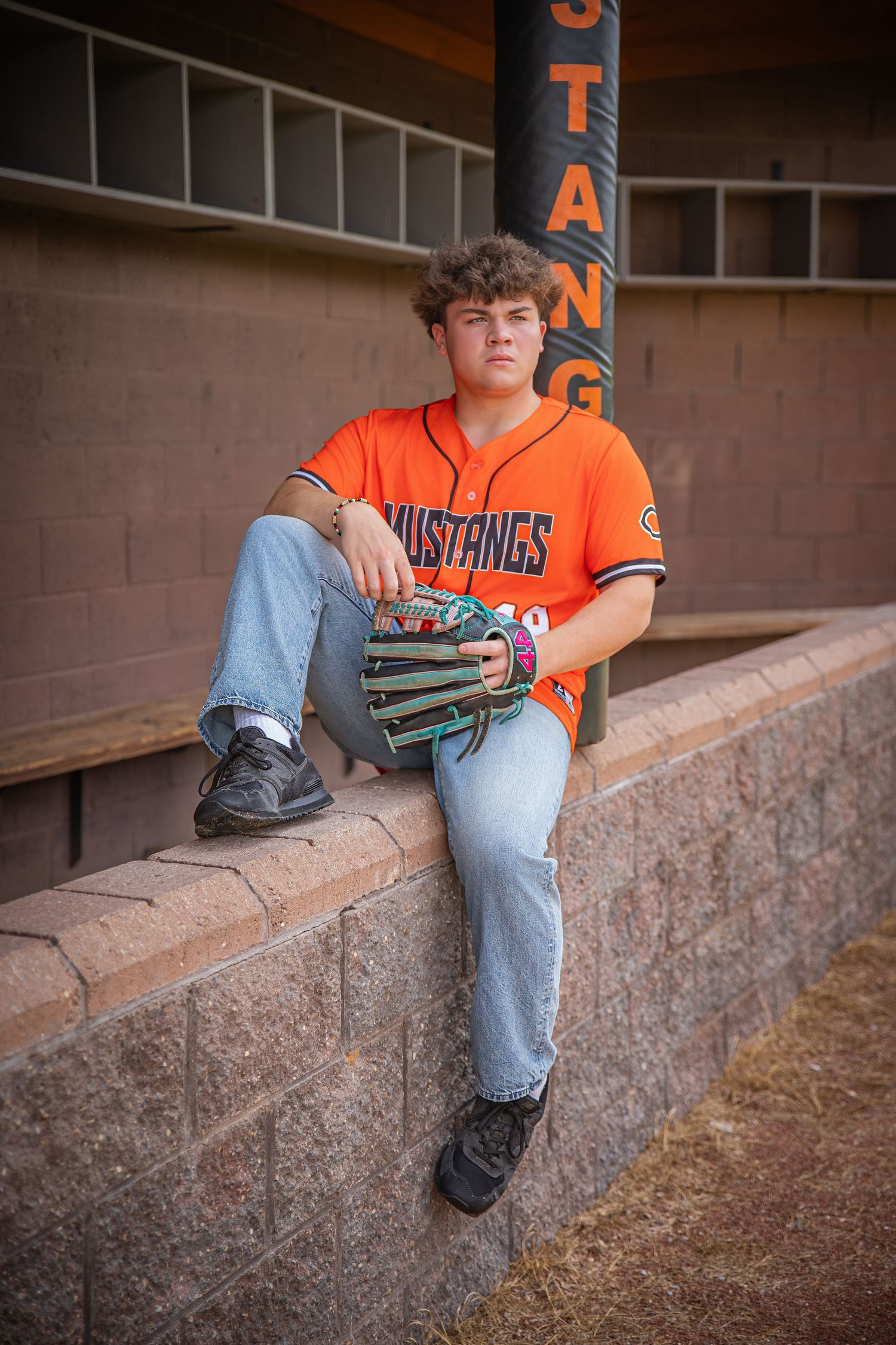 A baseball player in orange jersey sits on stadium wall with glove.