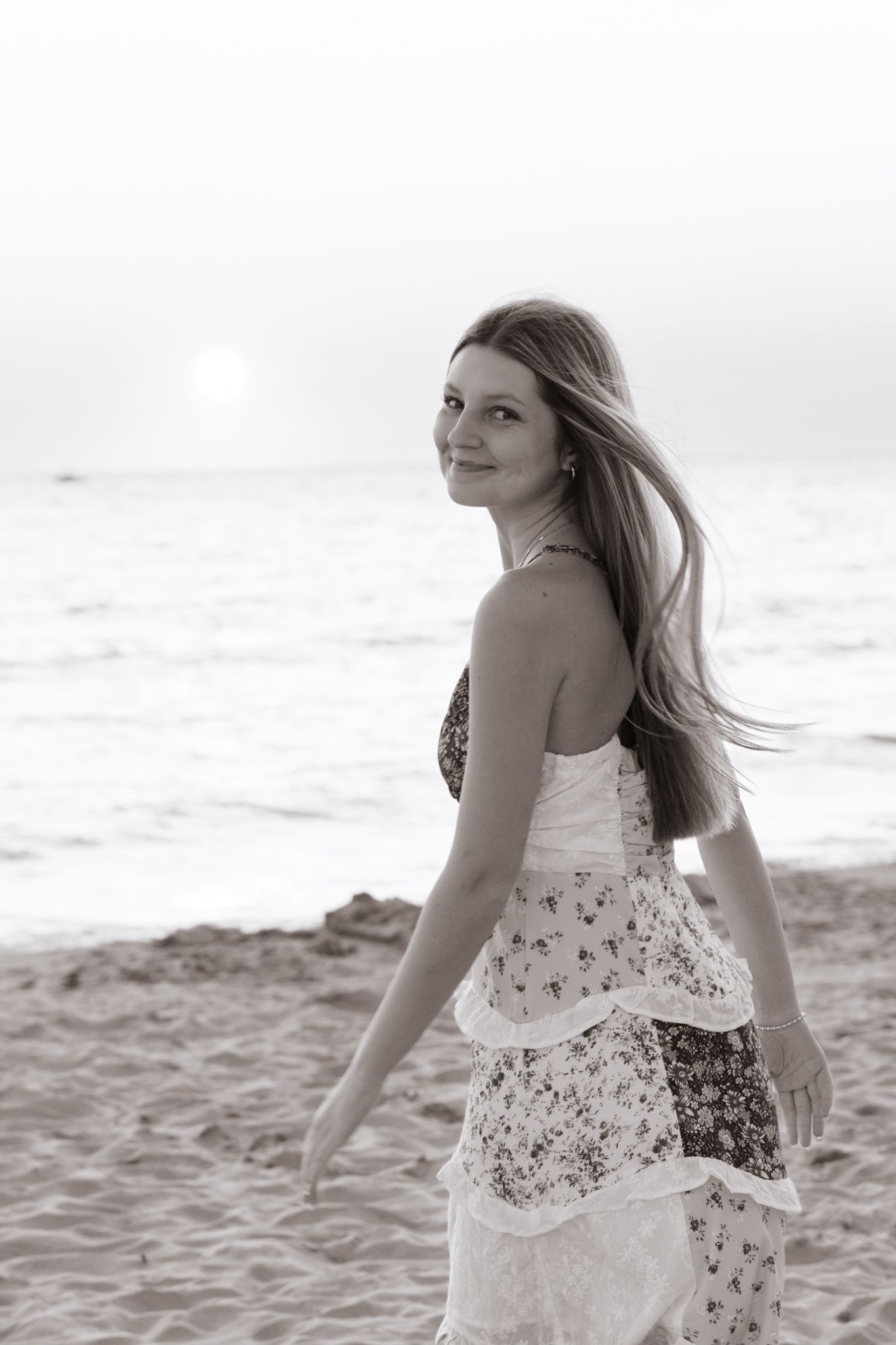 Black and white photo of a smiling woman in a floral dress walking on a sandy beach at sunset.