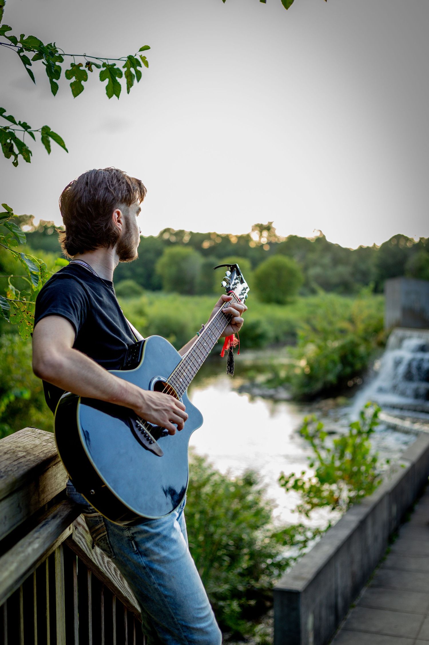 A musician with a blue acoustic guitar poses on a bridge overlooking a waterfall.