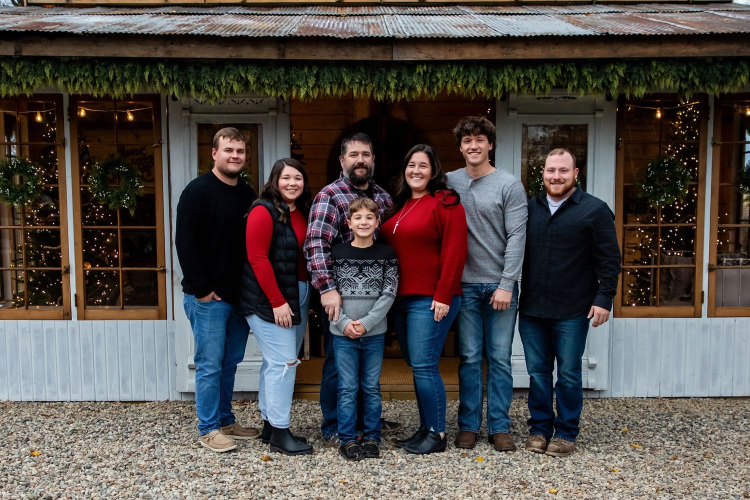 Family portrait session in casual attire standing outside a rustic building on a gravel path.